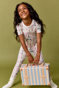 A Girl Wearing Fleur De La Tour Short Sleeve Stretch Jersey Kids Pj Set in a front view pose while holding a pastel suitcase 