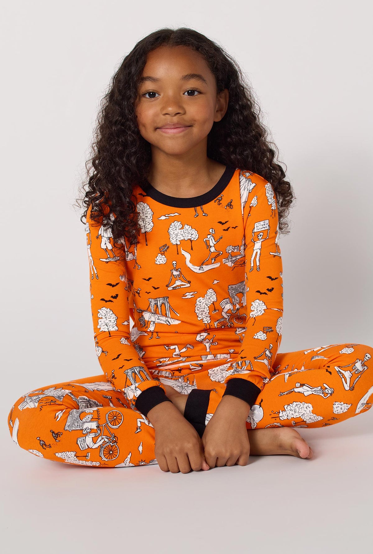A Girl Wearing Orange Spooky In The City Long Sleeve Stretch Jersey Kids Pj Set in a close up sitting pose