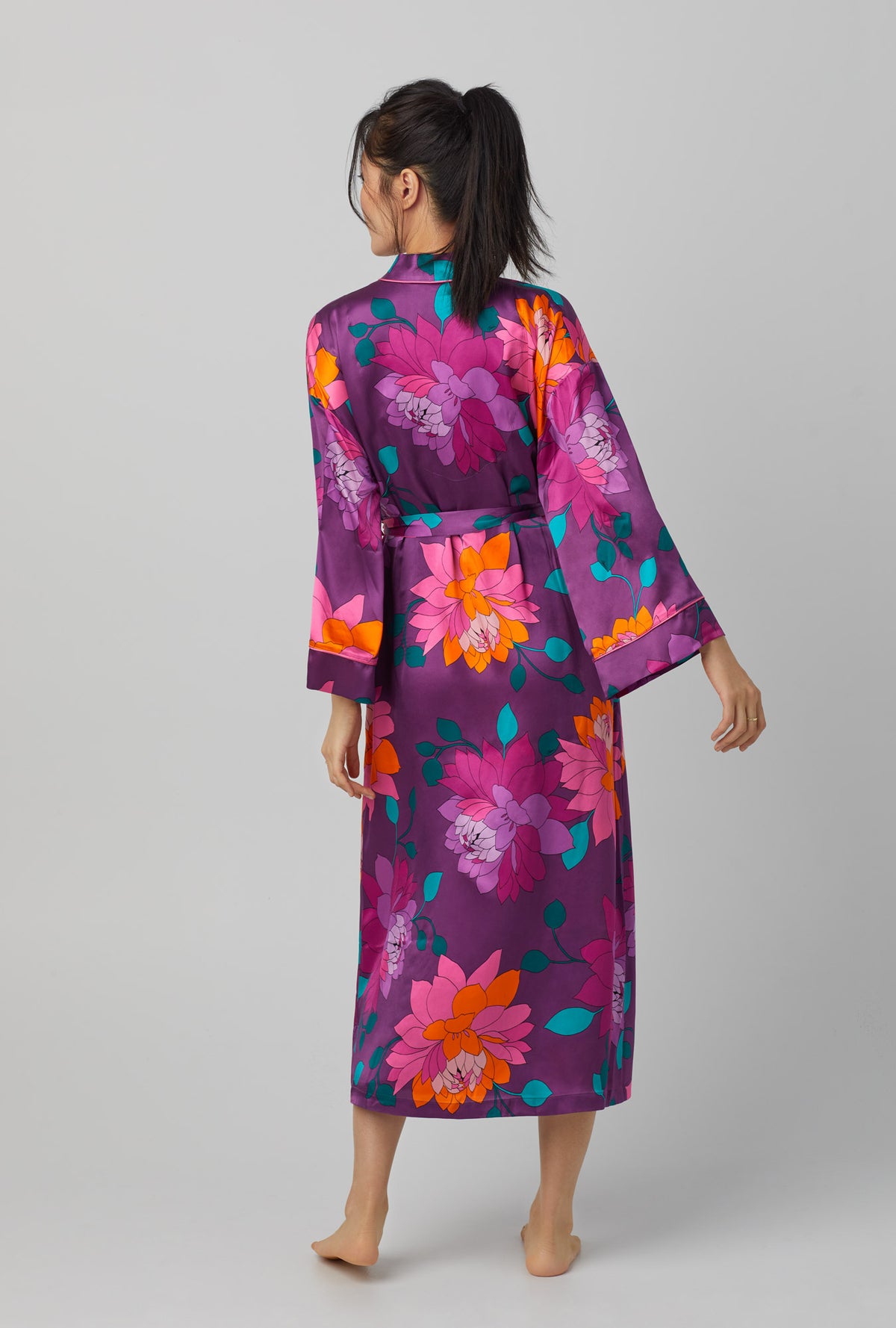 A lady wearing purple long sleeve banded collar washable silk maxi robe with trina turk evening blooms print.